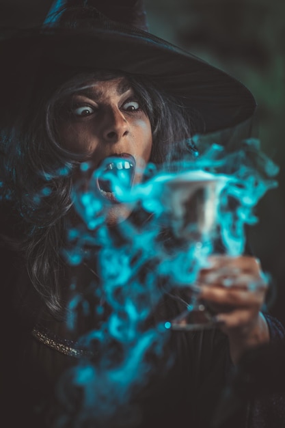 Porträt einer Hexe mit schrecklichem Gesicht in gruseliger Umgebung und rauchgrünem Hintergrund trinkt Zaubertrank aus dem Kelch.
