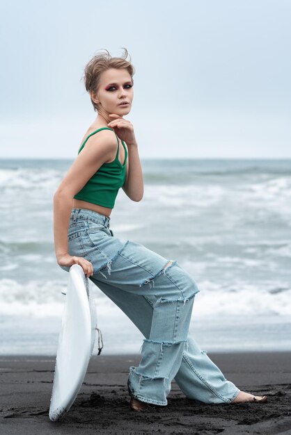 Foto porträt einer hipster-frau-surferin, die auf einem surfbrett sitzt und auf sand auf dem hintergrund von meereswellen liegt