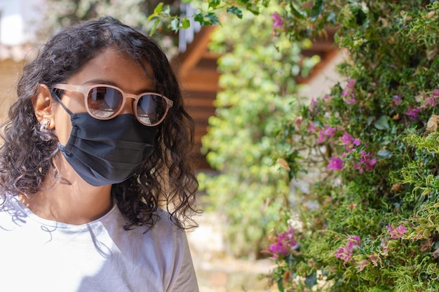 Porträt einer hispanischen Frau mit stilvoller Schutzmaske und Sonnenbrille, die auf natürlichem Hintergrund posiert