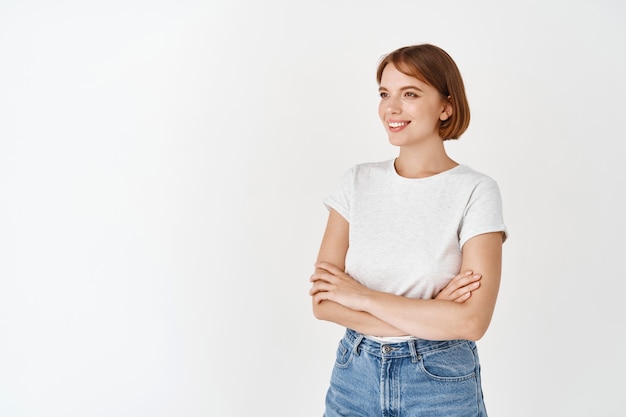 Porträt einer hoffnungsvollen lächelnden Frau im T-Shirt, die Arme auf der Brust verschränken mit glücklichem, fröhlichem Gesicht, weiße Wand