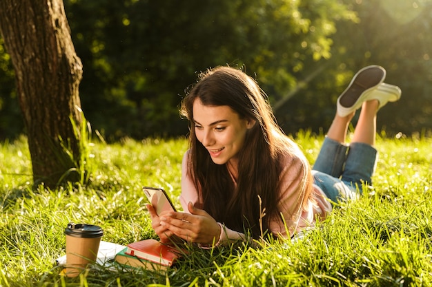 Porträt einer hübschen brünetten Frau, die an einem sonnigen Tag im Gras im grünen Park lächelt und Handy benutzt