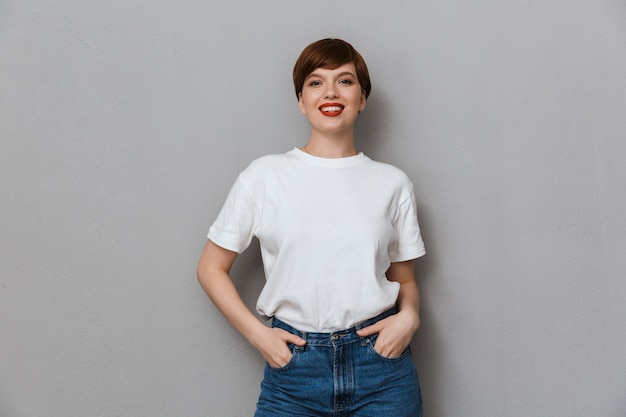 Porträt einer hübschen brünetten Frau, die ein lässiges T-Shirt trägt und vorne über grauer Wand isoliert lächelt