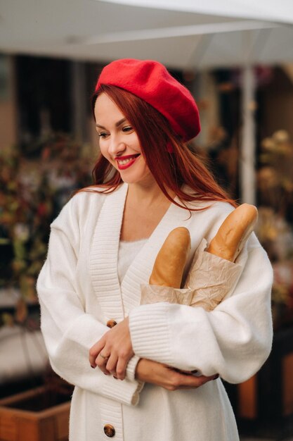 Porträt einer hübschen Frau in roter Baskenmütze und weißer Strickjacke mit Baguettes in den Händen, die durch die herbstliche Stadt schlendert