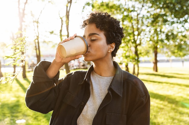 Porträt einer hübschen Frau mit lockigem Haar, die Kaffee zum Mitnehmen aus Pappbecher hält und trinkt, während sie im Stadtpark spazieren geht