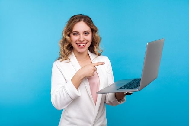 Porträt einer hübschen Geschäftsfrau mit welligem Haar in weißer Jacke, die auf einen Laptop zeigt und aufrichtig in die Kamera lächelt, zufrieden mit Anwendung, Computertechnologien. Studioaufnahme, blauer Hintergrund