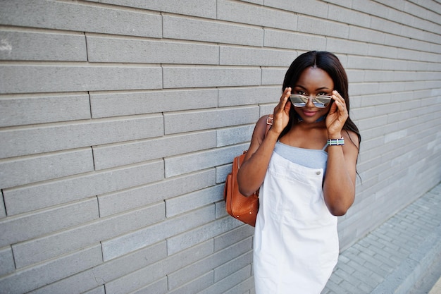 Porträt einer hübschen jungen Afroamerikanerin, die mit Sonnenbrille vor einer Backsteinmauer im Hintergrund posiert