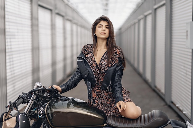 Porträt einer hübschen jungen Dame in Lederjacke und Kleid, die neben einem schwarzen Motorrad posiert und vor dem Hintergrund weißer Wände direkt in die Kamera schaut. Sexualitätskonzept