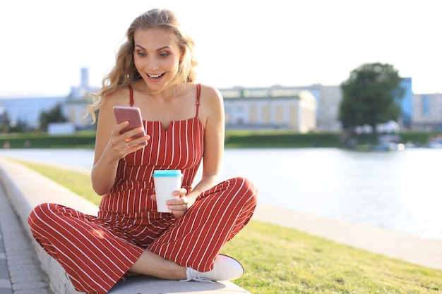 Porträt einer hübschen jungen Frau, die am Flussufer mit gekreuzten Beinen während des Sommertages sitzt, lächelt und Smartphone verwendet.