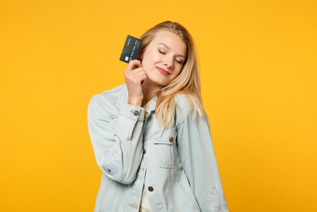 Porträt einer hübschen jungen Frau in Denim-Freizeitkleidung, die die Augen geschlossen hält und die Kreditkarte isoliert auf gelb-orangefarbenem Wandhintergrund im Studio hält. People-Lifestyle-Konzept. Mock-up-Kopienbereich.