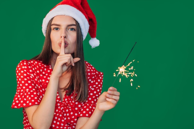 Porträt einer hübschen jungen Frau in einem weihnachtlichen roten Hut macht eine Geste des Schweigens Kleides in der Kleidung hält eine Wunderkerze in der Hand