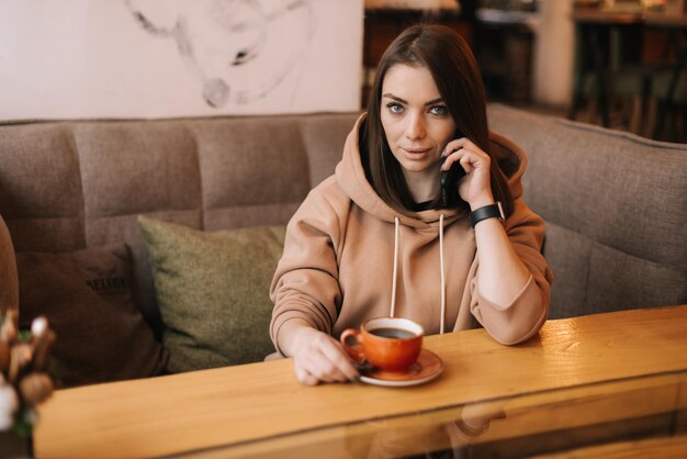 Porträt einer hübschen jungen Frau in Freizeitkleidung, die an einem Tisch in einem gemütlichen Café telefoniert. Kaffeetasse auf dem Tisch. Mädchen, das die Kamera betrachtet. Konzept der Freizeitbeschäftigung.