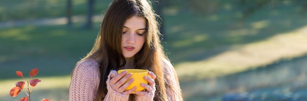 Porträt einer hübschen jungen Frau in stylischem Strickpullover, die an einem sonnigen Tag auf dem goldenen Herbstwald steht Zeit zum Entspannen und einen Kaffee trinken