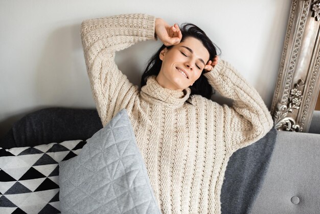 Porträt einer hübschen jungen kaukasischen Frau, die auf einem grauen Sofa in der Nähe des Weihnachtsbaums sitzt und ein Kissen hält und mit geschlossenen Augen mit einem schönen Lächeln aufwacht Weihnachts- und Neujahrsstimmung