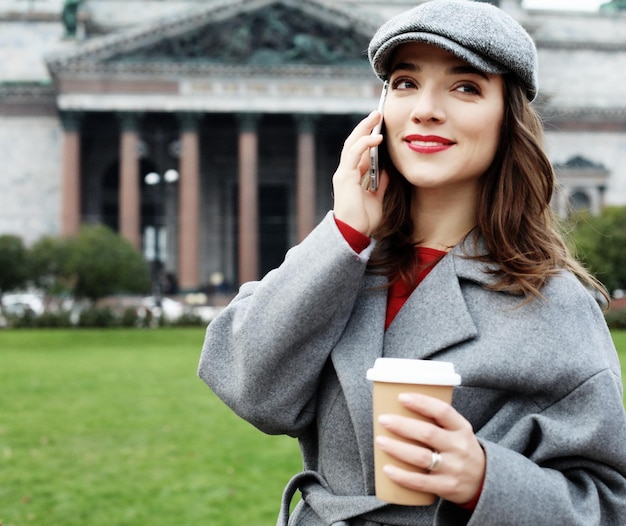 Porträt einer hübschen lächelnden Frau unter Verwendung des Mobiltelefons beim Halten der Kaffeetasse auf einer Stadtstraße