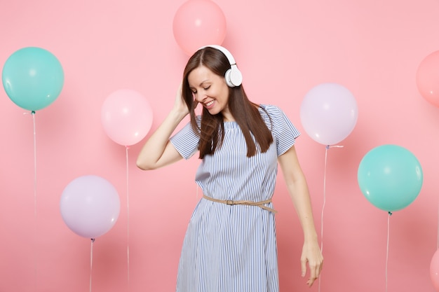 Porträt einer hübschen lächelnden jungen Frau mit geschlossenen Augen mit Kopfhörern im blauen Kleid, die Musik auf rosafarbenem Hintergrund mit bunten Luftballons hört. Geburtstagsfeier-Partyleute aufrichtige Gefühle.