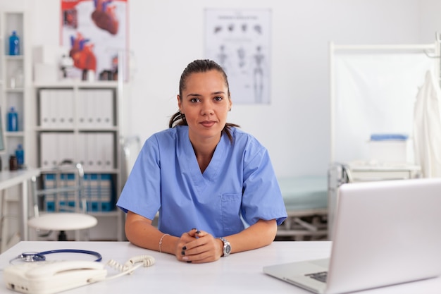 Porträt einer hübschen medizinischen Krankenschwester, die vorne im Krankenhausbüro lächelt und blaue Uniform trägt