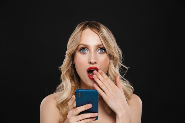 Porträt einer hübschen schockierten jungen blonden Frau mit den roten Lippen des hellen Make-ups, die lokalisiert unter Verwendung des Handys aufwerfen.