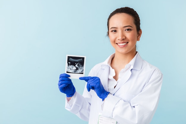 Porträt einer jungen ärztin, die isoliert über der blauen wand posiert, die röntgenaufnahme des babys in den händen hält.