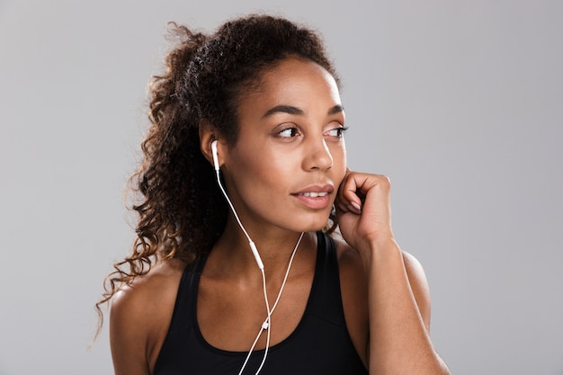 Porträt einer jungen afroamerikanischen jungen Sportlerin lokalisiert über grauem Hintergrund, Musik mit Kopfhörern hörend, weg schauend