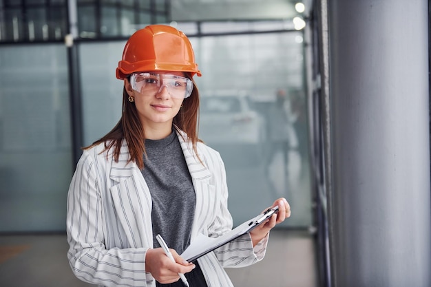 Porträt einer jungen Arbeiterin mit Schutzhelm, Brille und Notizblock. Stehen im Büro.