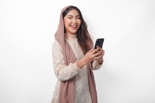 Porträt einer jungen asiatischen muslimischen Frau im Schleier-Hijab, die fröhlich lächelt, während sie ein Smartphone hält und verwendet, Ramadan und Eid Mubarak Konzept