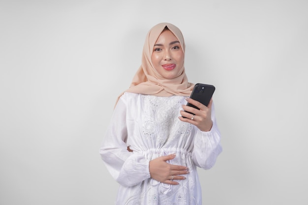 Foto porträt einer jungen asiatischen muslimischen frau in weißem kleid und hijab, die sich hungrig fühlt, während sie auf ihr handy schaut, um nach dem fasten leckeres essen zu bestellen, isoliert auf weißem hintergrund