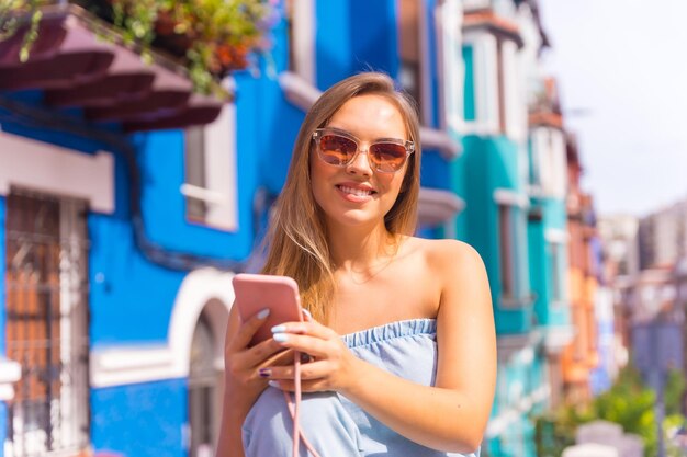 Porträt einer jungen attraktiven blonden Frau mit Sonnenbrille, die hinter einer blauen, farbenfrohen Fassade auf das Telefon blickt