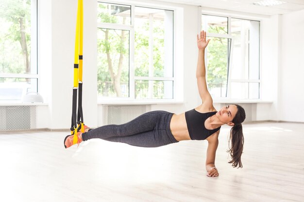 Porträt einer jungen attraktiven muskulösen Frau in schwarzem Oberteil und Leggings, die auf perfekter Seitenplankenposition steht, verwendet Fitnessgurte und macht Liegestütze, während die Beine am TRX hängen. Innen, Fensterhintergrund