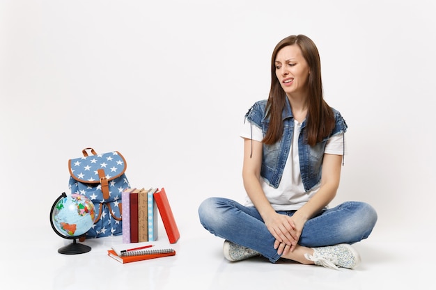 Porträt einer jungen besorgten verwirrten Studentin in Denim-Kleidung, die isoliert auf Globusrucksackschulbüchern sitzt und schaut