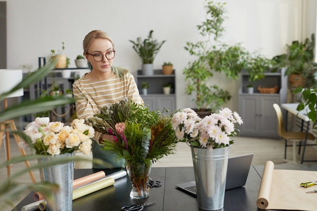 Porträt einer jungen blonden Frau, die Blumensträuße arrangiert, während sie in der grünen Floristenwerkstatt arbeitet, Kopierraum