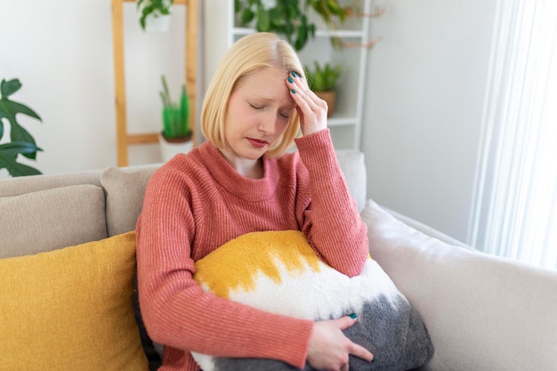 Porträt einer jungen blonden Frau, die zu Hause mit Kopfschmerzen und Migräne auf der Couch sitzt. Schöne Frau, die unter chronischen täglichen Kopfschmerzen leidet. Traurige Frau, die ihren Kopf wegen Nebenhöhlenschmerzen hält