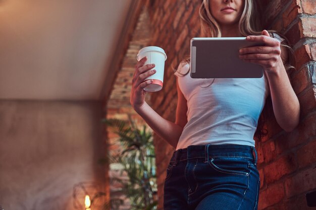 Porträt einer jungen blonden Frau hält eine Tasse Kaffee zum Mitnehmen und benutzt ein digitales Tablet, während sie sich in einem Raum mit Loft-Interieur an eine Ziegelwand lehnt.