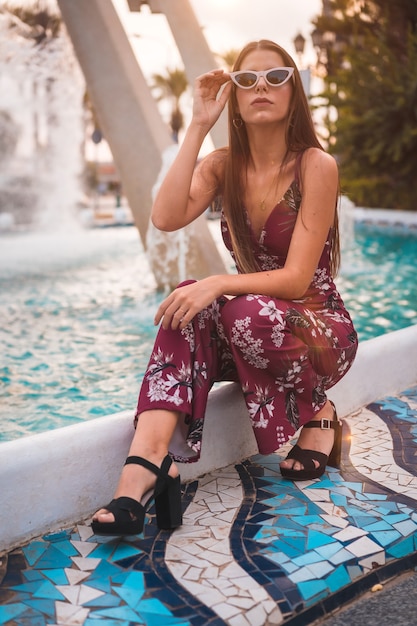 Porträt einer jungen Brünette in einem kastanienbraunen Blumenkleid, das Sommer in der goldenen Stunde durch einen Stadtbrunnen genießt