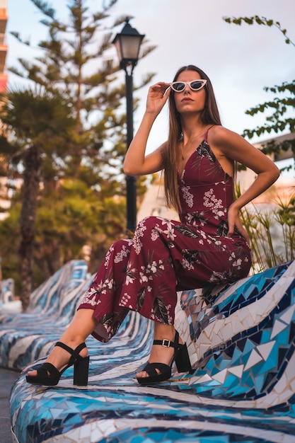 Porträt einer jungen Brünette in einem kastanienbraunen Blumenkleid, das Sommer in der goldenen Stunde durch einen Stadtbrunnen genießt