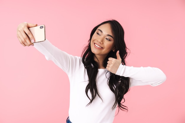 Porträt einer jungen brünetten Asiatin mit langen Haaren, die lächelt und ein Selfie-Foto auf dem Handy macht, isoliert über rosa Wand