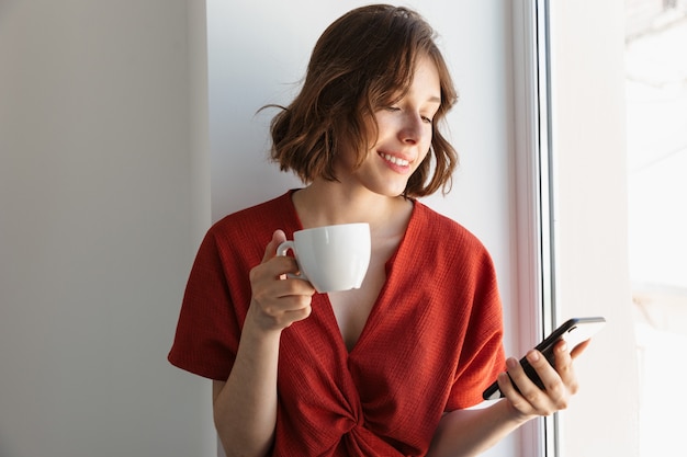 Porträt einer jungen brünetten Frau in Freizeitkleidung, die Kaffee trinkt und Smartphone benutzt, während sie zu Hause über dem Fenster sitzt