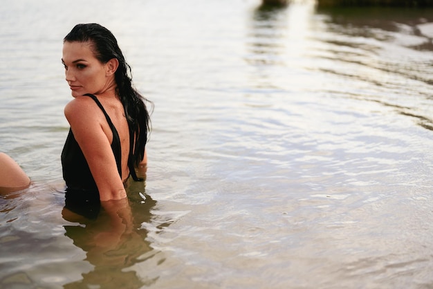 Porträt einer jungen brünetten Frau sitzt im Wasser auf See. Mädchen im schwarzen Badeanzug genießt den Sommer. Platz kopieren.