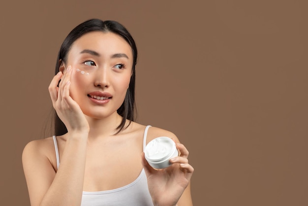 Porträt einer jungen chinesischen Dame mit Feuchtigkeitscreme auf ihrem hübschen Gesicht, die ein neues Schönheitsprodukt testet