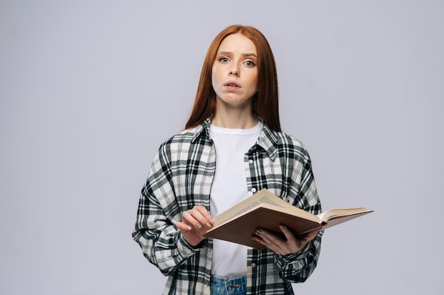 Porträt einer jungen College-Studentin, die geöffnete Bücher hält und auf isoliertem grauem Hintergrund in die Kamera blickt. Hübsches rothaariges Damenmodell, das legere Kleidung trägt und emotional Gesichtsausdrücke zeigt