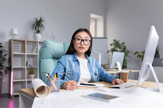 Porträt einer jungen Designerin im modernen Büro bei der Arbeit erfolgreiche asiatische Frau mit Brille, die in die Kamera blickt