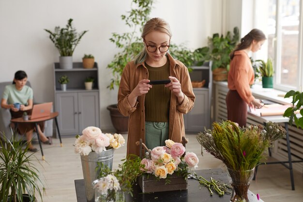 Foto porträt einer jungen floristin, die fotos von blumenkompositionen über das smartphone macht, während sie im blumenladen arbeitet, platz kopieren