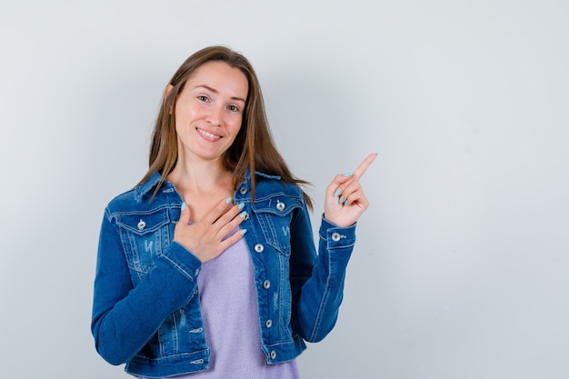 Porträt einer jungen Frau, die auf die obere rechte Ecke zeigt, mit der Hand auf der Brust in T-Shirt, Jacke und dankbarer Vorderansicht