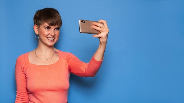 Porträt einer jungen Frau, die ein Selfie auf einem Smartphone auf blauem Hintergrund macht. Ein Ort für Ihren Text. Das Konzept der Videokommunikation auf Distanz.