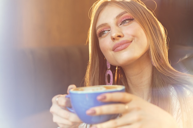 Porträt einer jungen Frau, die eine Tasse Kaffee trinkt und durch das Fenster schaut