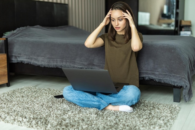 Porträt einer jungen Frau, die frustriert und verzweifelt, wütend und traurig auf dem Boden sitzt, mit den Händen auf dem Kopf. Halten eines Laptops.