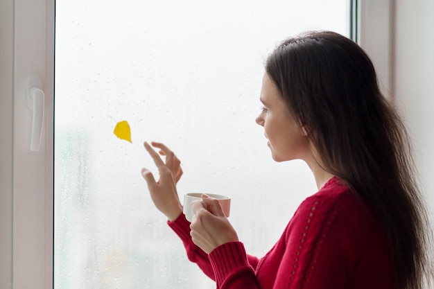 Porträt einer jungen Frau, die mit Regentropfen aus dem Fenster schaut, gelbes Blatt im Herbst. Frau, die eine Tasse Kaffee genießt