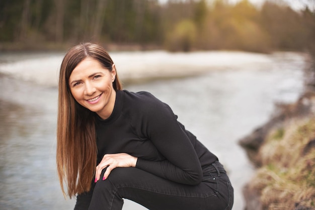 Porträt einer jungen Frau, die schwarze Jeans und ein Oberteil trägt, sich lehnt, ihr langes Haar fällt herunter und lächelt. Verschwommener Fluss im Hintergrund