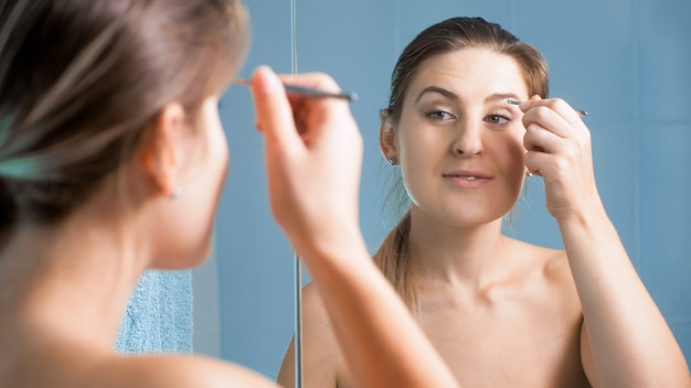 Porträt einer jungen Frau, die sich mit einer Pinzette im Badezimmer um ihre Augenbrauen kümmert.