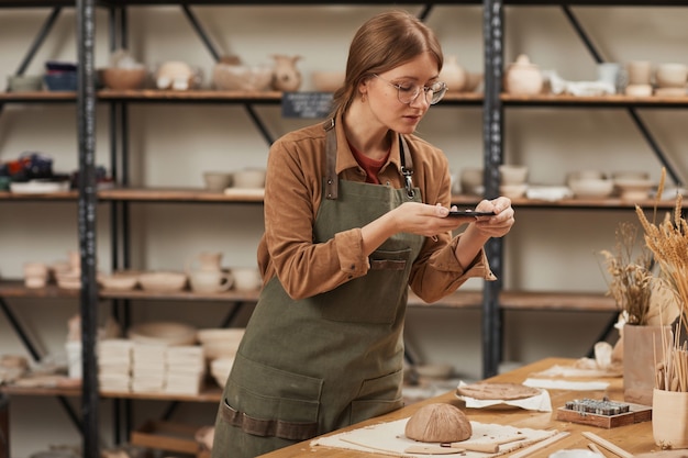 Porträt einer jungen Frau, die Smartphone-Foto von roher Keramikschale in der Töpferwerkstatt macht, um sie in sozialen Medien zu veröffentlichen, Konzept für kleine Unternehmen, Kopierraum