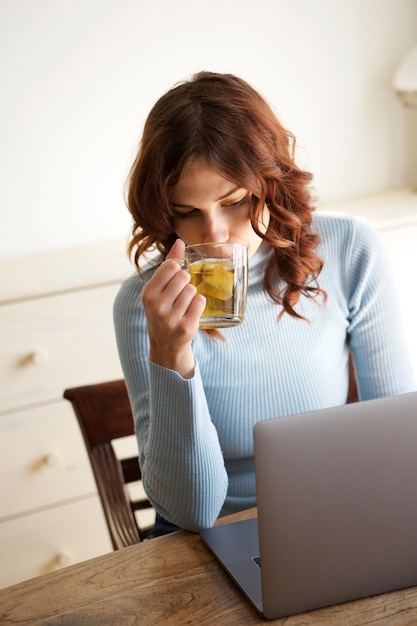 Porträt einer jungen Frau, die Tee trinkt, während sie am Laptop arbeitet